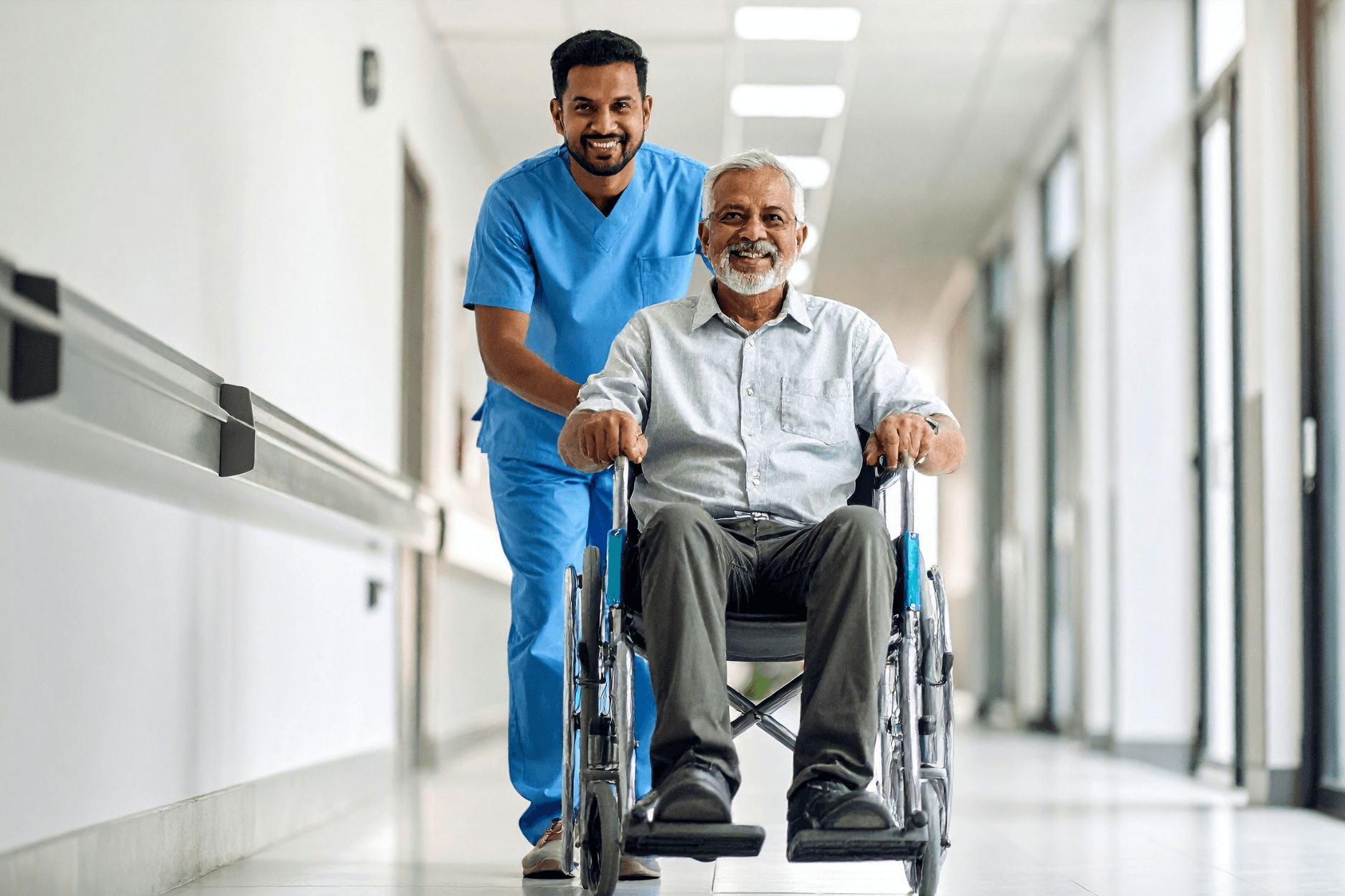 A smiling male caregiver pushing an elderly man in a wheelchair through a bright corridor